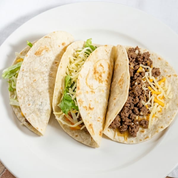 Three soft tacos filled with ground beef, shredded cheese, and lettuce on a white plate.