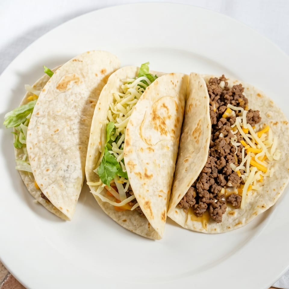 Three soft tacos filled with ground beef, shredded cheese, and lettuce on a white plate.