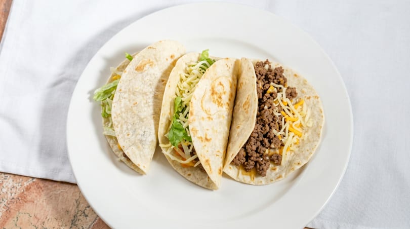 Three soft tacos filled with ground beef, shredded cheese, and lettuce on a white plate.